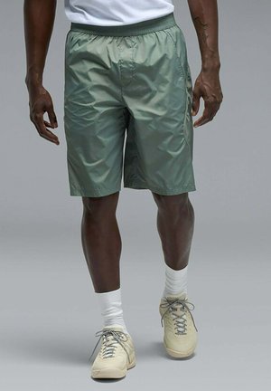 Uomo che indossa pantaloncini verde chiaro fino al ginocchio, calze bianche, sneakers beige e una camicia bianca, in piedi su uno sfondo grigio.