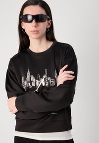 Sweatshirt preto com um gráfico do horizonte da cidade, gola redonda e bainha canelada. Usado com óculos de sol pretos e brincos de argola prateados.