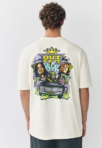 Kermainen puuvillainen t-paita, jossa on värikäs graafinen kuvio ja rohkeat tekstit. Kuvassa on kaksi hahmoa, auto ja kruunu paidan takana.