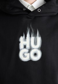 Sort hættetrøje med hætte, prydet med ordet "HUGO" i fede hvide bogstaver med en røgfyldt effekt, sat imod en glat stofstruktur.