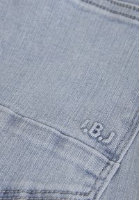 Ljusblå denim med en slät textur, prydd med sömdetaljer och en broderad logotyp i det nedre hörnet av fickan.