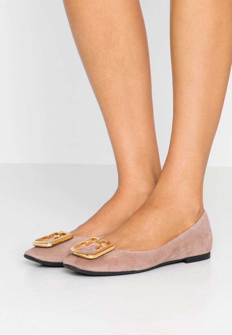 Jambes portant des chaussures plates en daim mauve avec de grands accents de boucles rectangulaires dorées sur un sol blanc et un fond gris.