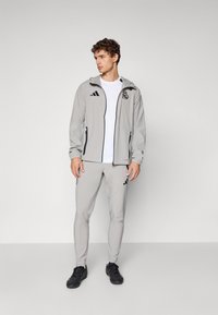 Joven de pie vistiendo un chándal gris claro del Real Madrid con logotipos negros de Adidas y zapatillas deportivas negras, contra un fondo liso.