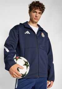 Marineblaues Zip-Jackett mit weißen Akzenten und Real Madrid-Logo, kombiniert mit einem Fußball, der schwarze und pinke Sternmuster zeigt.