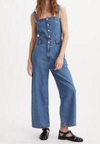 Kvinna i ärmlös blå denim jumpsuit med framknappar och svarta öppna sandaler, stående mot en enkel vit bakgrund.