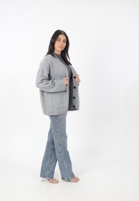 Cardigan grigio a maglia con un fit ampia, grandi tasche frontali e cinque bottoni neri, indossato sopra un top aderente e abbinato a jeans a zampa.