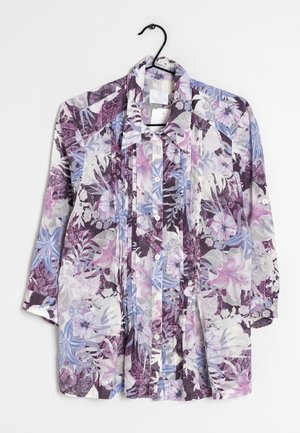 Langærmet skjorte med knapper og print med lilla, lyserøde og blå blomster og blade, hængende på en sort bøjle mod en hvid baggrund.