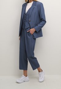 Niebieska, dopasowana marynarka na dopasowanej kamizelce i spodniach z wysokim stanem. Gładka tkanina, zapinana na dwa guziki, z bocznymi kieszeniami. W połączeniu z białymi trampkami.