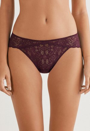 Vrouw in paarse kanten hipsteronderbroek, zichtbaar vanaf de taille tot de bovenbenen met handen ontspannen naast zich.