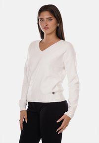 Maglione bianco a maglia con scollatura a V, orlo a coste e maniche lunghe. Presenta una piccola accentuazione metallica rotonda sul lato. Tessuto morbido.