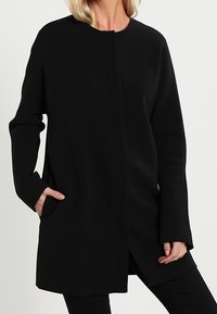 Černý dlouhý kabát s rovný střihem, kulatým výstřihem, bočními kapsami a hladkou strukturou. Nemá viditelné zapínání ani vzory.