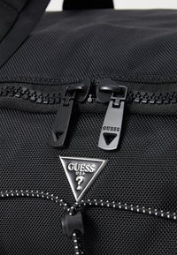 Sac à dos noir en nylon avec des zips noirs double, une étiquette de logo triangulaire et un accent en cordon élastique avec du fil blanc.
