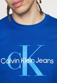 Camiseta de algodón azul con "Calvin Klein Jeans" en texto blanco y azul claro, cuello redondo y mangas cortas. Textura suave y lisa.