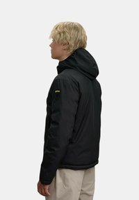 Schwarze isolierte Jacke mit Kapuze, mit einem Logo-Patch am Oberarm. Glatte Oberfläche, durchgehender Reißverschluss und Seitentaschen.