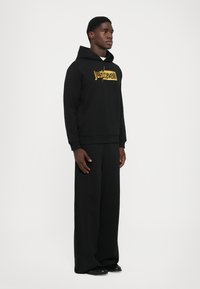 Felpa nera con cappuccio realizzata in tessuto morbido, con un logo dorato "Just Cavalli" centrato. Abbinata a pantaloni neri larghi e scarpe nere.