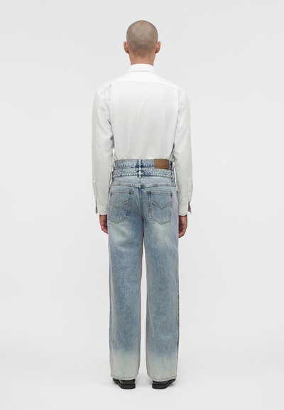 Άνδρας με λευκό πουκάμισο μακρυμάνικο και ξεθωριασμένο μπλε τζιν με ψηλή μέση και φαρδιά πόδια, που στέκεται με την πλάτη προς τα εμπρός σε μονόχρωμο φόντο.