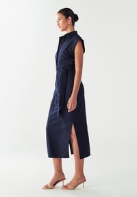 Abito in lino blu navy con maniche corte, chiusura a bottoni frontale, vita addolcita da una cintura e spacchi laterali. Abbinato a sandali con tacco di un beige chiaro.