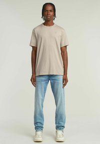 Maglietta beige a maniche corte con un logo sottile, abbinata a jeans in denim azzurro chiaro. Il tessuto sembra morbido e presenta una vestibilità rilassata.