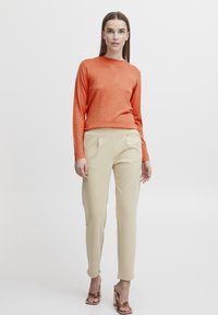 Pulover lung tricotat de culoare portocalie, cu mâneci lungi, asortat cu pantaloni cremoși, cu croială tapered, având pliseuri și o textură netedă. Modelul poartă sandale maro cu vârf deschis.
