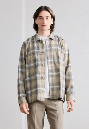 Νέος άνδρας με μεσαίου μήκους σκούρα μαλλιά, φορώντας ένα μπεζ και μαύρο καρό πουκάμισο πάνω από ένα λευκό T-shirt και μπεζ παντελόνια, στέκεται ουδέτερα.