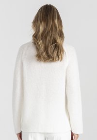 Kobieta z długimi falującymi, jasnobrązowymi włosami, ubrana w biały puszysty sweter i białe spodnie, stojąca tyłem do kamery na szarym tle.