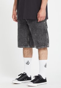 Shorts en denim noir cargo avec une finition délavée, dotés de poches avant et d'une poche à rabat sur le côté, associés à des baskets noires et des chaussettes blanches avec logo.