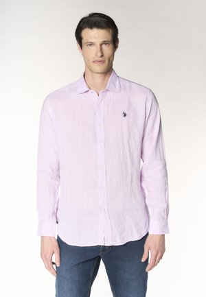 Uomo che indossa una camicia rosa chiaro a manica lunga con bottoni e logo ricamato, abbinata a jeans blu, in piedi su uno sfondo semplice.