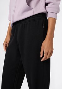 Pantalones de chándal negros hechos de una tela suave, con una cintura elástica y una pequeña etiqueta de marca en el costado, combinados con una sudadera lila.