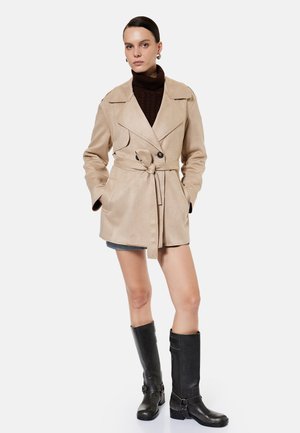 Frau trägt beigen Trenchcoat mit Gürtel, dunkelbraunen Rollkragenpullover, kurzen Rock und schwarze kniehohe Stiefel, steht mit den Händen in den Taschen.