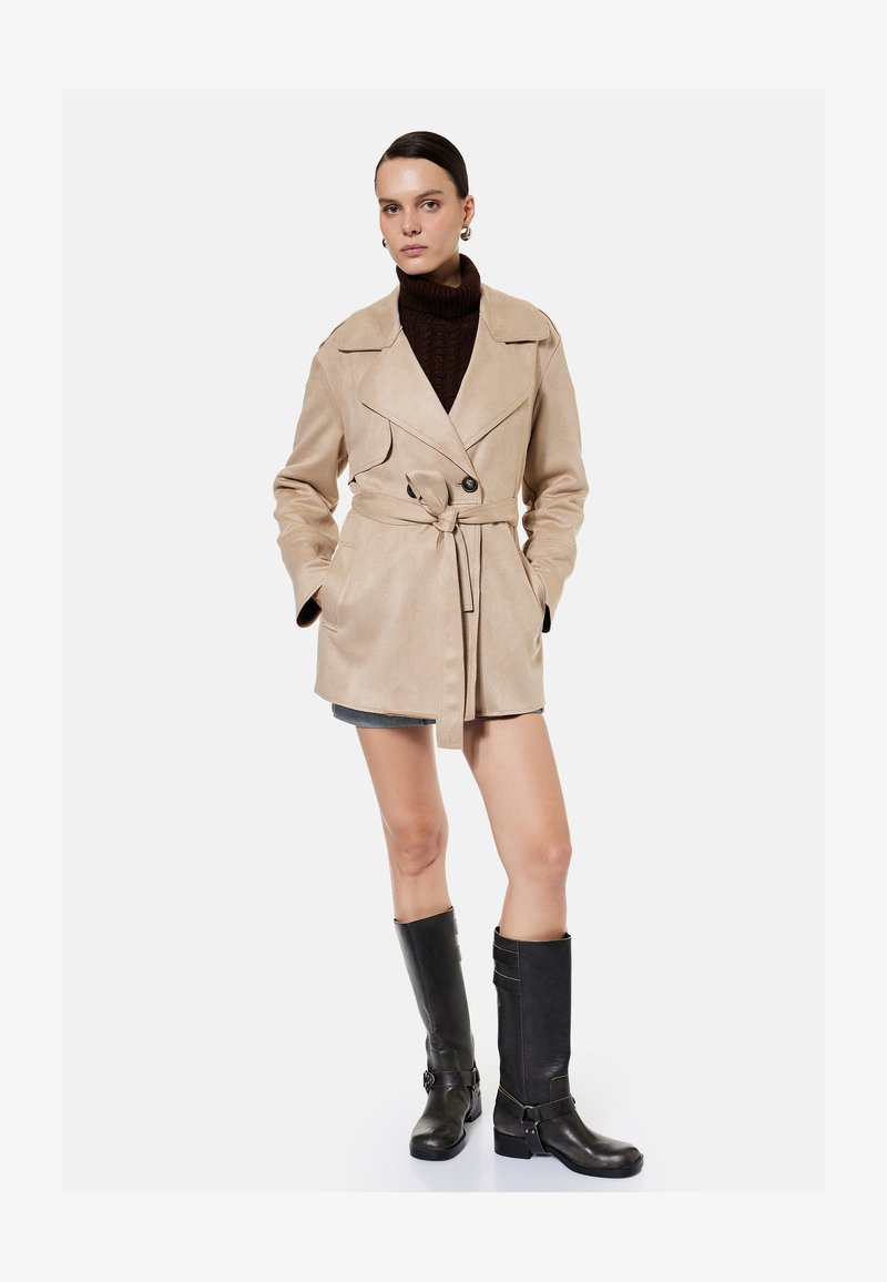 Kvinde iført beige trenchcoat med bælte, mørkebrun rullekrave, kort nederdel og sorte knælange støvler, stående med hænderne i lommerne.
