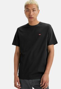 Ung mand med kort blond hår iført en sort Levi's t-shirt og grå jeans, stående mod en ensfarvet hvid baggrund.