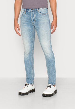 Uomo che indossa jeans slim fit azzurri chiaro e scarpe bianche con lacci, in piedi contro uno sfondo chiaro e semplice.