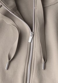 Felpa con zip beige, con una texture liscia, dotata di una cerniera argentata, cordini e un cappuccio accogliente. Design semplice e minimale.
