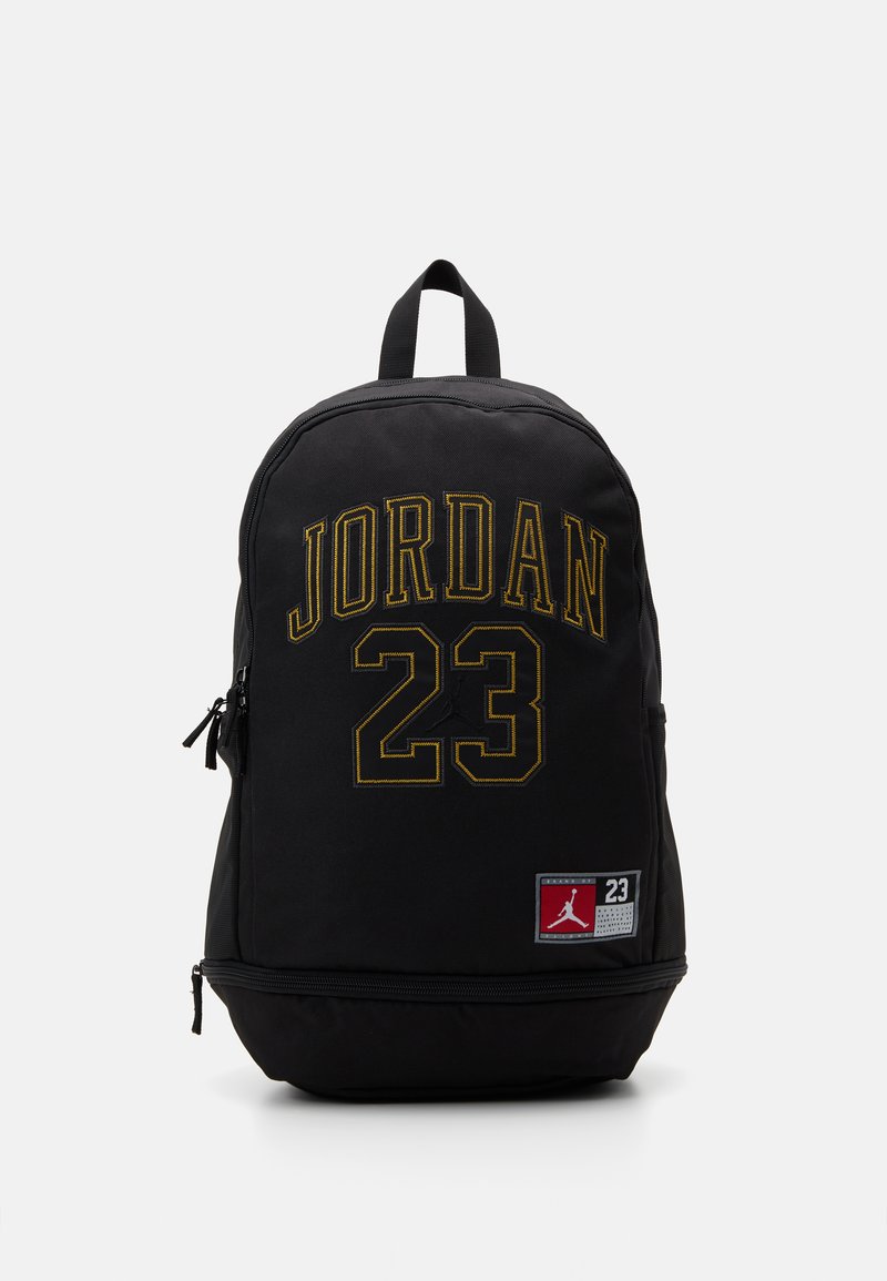 Mochila negra con texto amarillo "JORDAN" y "23" en la parte frontal, bolsillos con cierre, tela duradera, forma redondeada y correas de hombro acolchadas.