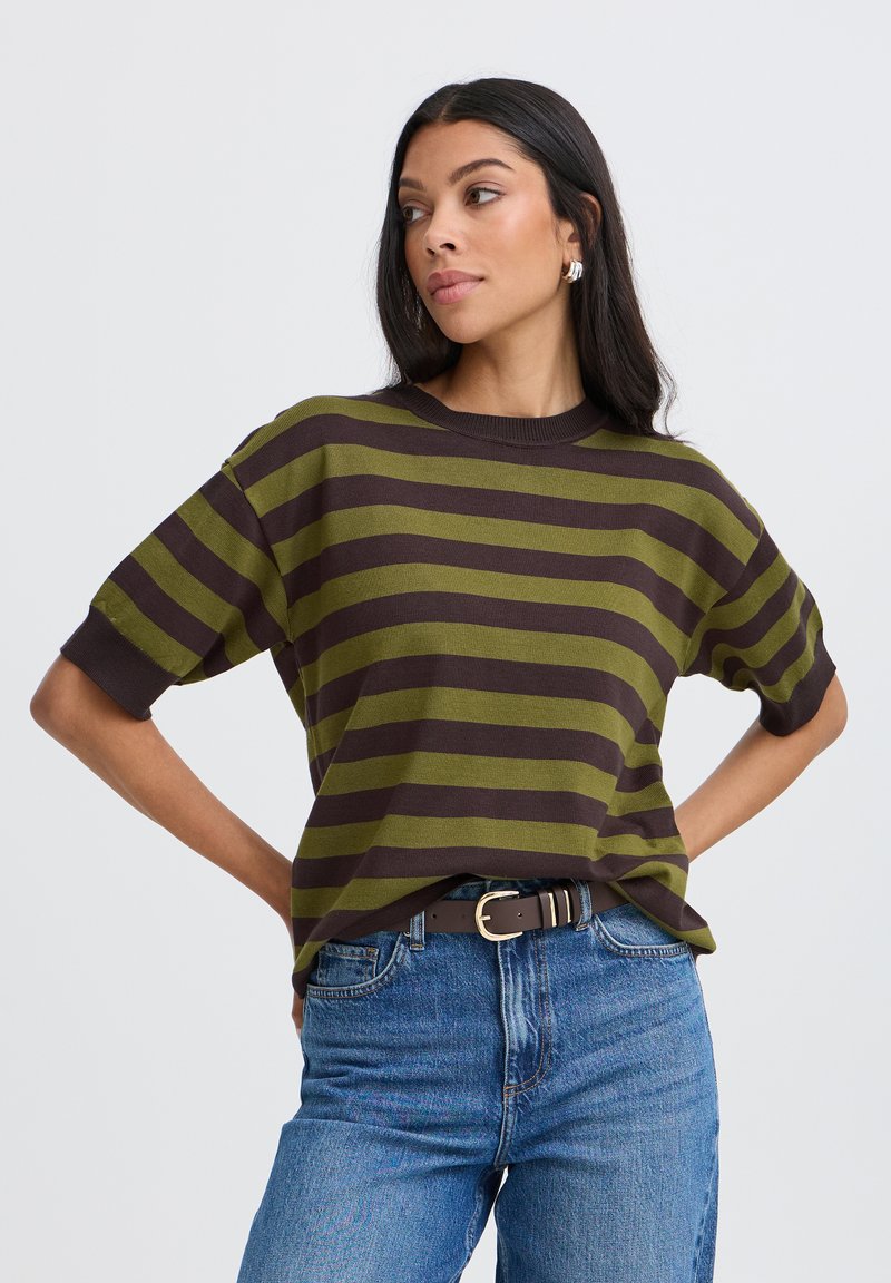 Femme aux longs cheveux foncés portant un t-shirt à manches courtes rayé vert et marron, rentré dans un jean bleu avec une ceinture noire, regardant sur le côté.
