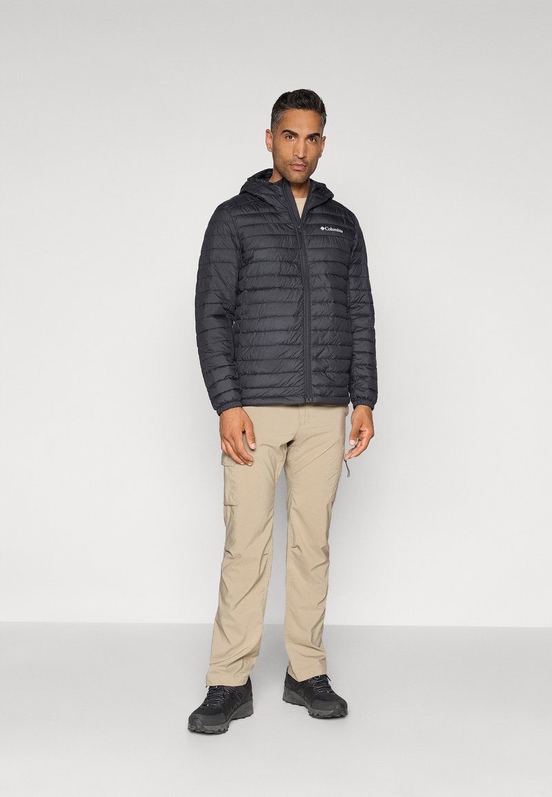 Chaqueta negra acolchada con capucha, que presenta un logo en el pecho, combinada con pantalones cargo ligeros beige y zapatos de senderismo negros.