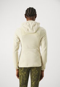 Chaqueta de forro polar beige claro con capucha, de superficie texturizada, diseño ajustado y costuras visibles; combinada con leggings verdes con patrones.