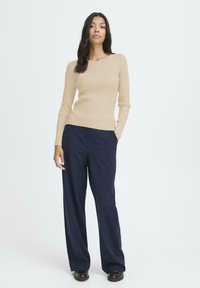 Donna con lunghi capelli scuri che indossa un maglione beige a coste, pantaloni a gamba larga blu notte e scarpe scure, in piedi con una mano in tasca.