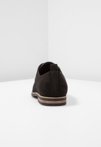 Chaussure en daim noir à forme arrondie, avec une semelle marron contrastante et des détails de coutures discrètes. Le talon est équipé d'une petite languette de tirage.