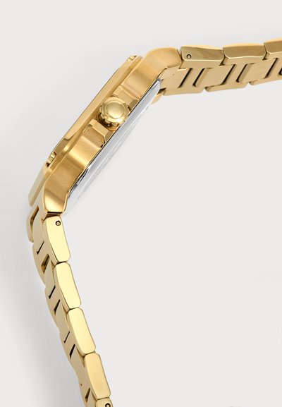 Gros plan d’un bracelet de montre en métal doré et du côté du boîtier de la montre avec une couronne sur un fond blanc uni.
