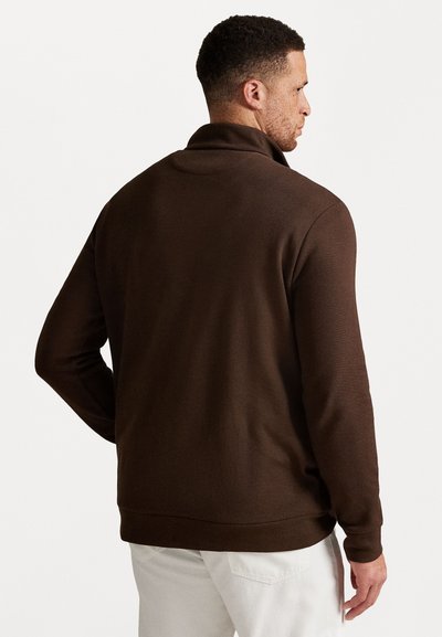 Bruine fleece jas met ritssluiting en hoge kraag, met een gladde textuur en lange mouwen, gedragen over lichte broek.