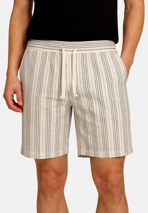 Hombre vistiendo pantalones cortos de color beige claro con finas rayas verticales negras y una cintura elástica con cordón, con las manos en los bolsillos.