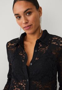 Schwarzes Spitzenhemd mit Knopfleiste, transparentem Stoff und floralem Muster. Lange Ärmel und klassischer Kragen vervollständigen den Look.