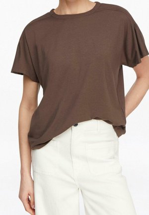 Personne portant un t-shirt marron à manches courtes ample, légèrement rentré dans un pantalon blanc taille haute avec poches avant.