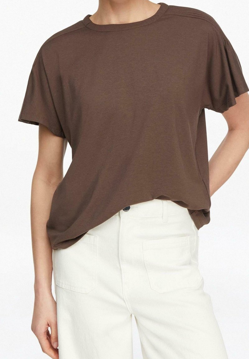 Personne portant un t-shirt marron à manches courtes ample, légèrement rentré dans un pantalon blanc taille haute avec poches avant.