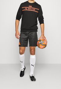 Felpa nera con logo dell'AC Milan e strisce arancioni, pantaloni corti grigi, calze bianche e scarpini neri. Tiene un pallone da calcio arancione a fantasia.
