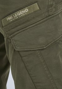 Poche de pantalon cargo vert olive avec rabat et étiquette en tissu PME Legend American Classic cousue sur la cuisse.
