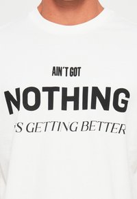 Person som har på seg en hvit skjorte med svart tekst som sier "AIN'T GOT NOTHING IS GETTING BETTER" på brystet.
