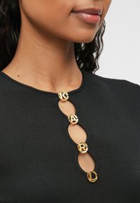 Svart tettsittende topp med korte ermer, med gyldne logo-knapper og nøkkelhullskutinger ved utringningen, teksturert ribbestrikket stoff.