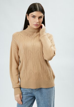 Beige gestrickter Pullover mit Stehkragen, aufwendigen Zopfmustern und gerippten Bündchen. Kombiniert mit hellblauen Jeans.